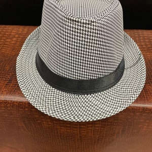 May include: A fedora hat with a black and white houndstooth pattern. The hat features a black band around the crown and a wide brim. The hat is a classic accessory.