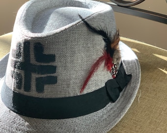 May include: A gray fedora hat with a black band and a black and white geometric design. The hat has a red and black feather accent.