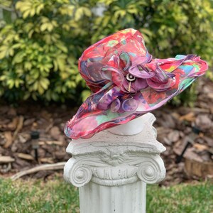 May include: A large, wide-brimmed hat with a colorful abstract print in shades of pink, red, green, and blue. The hat has a large, ruffled flower embellishment in shades of purple and pink.