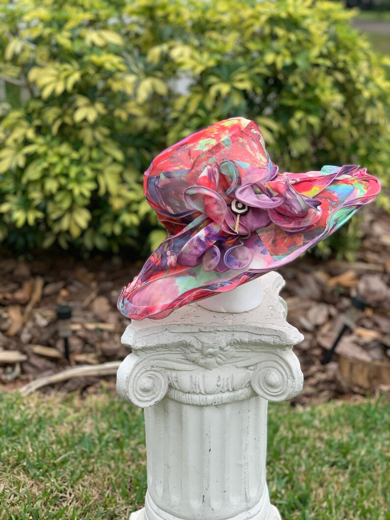 May include: A large, wide-brimmed hat with a colorful abstract print in shades of pink, red, green, and blue. The hat has a large, ruffled flower embellishment in shades of purple and pink.