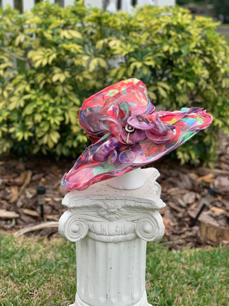 May include: A large, pink and purple floral print hat with a ruffled brim. The hat is sitting on a white pedestal.