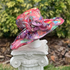 May include: A colorful, floral patterned hat with a large pink flower accent. The hat is made of a sheer fabric and is sitting on a white pedestal.