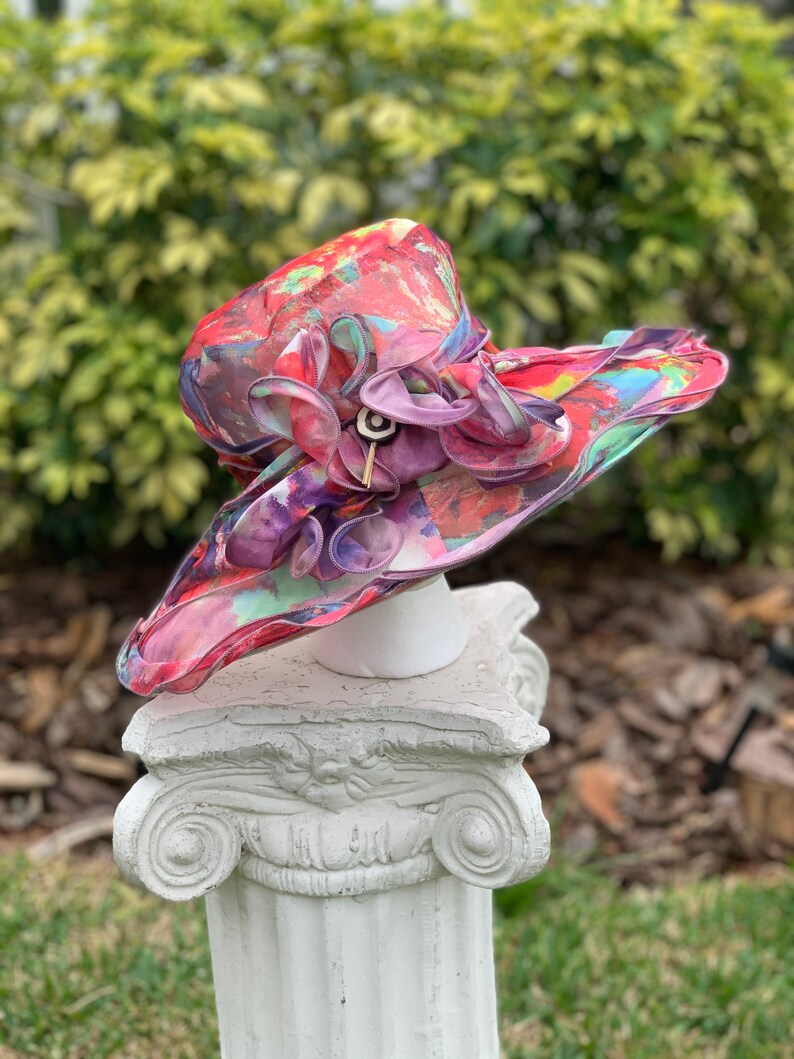 May include: A colorful, floral patterned hat with a large pink flower accent. The hat is made of a sheer fabric and is sitting on a white pedestal.
