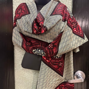 May include: A beige and red patterned scarf with a geometric design. The scarf is made of a soft, silky fabric and features a repeating pattern of circles and squares.