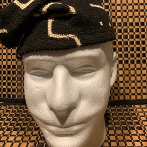 May include: A black and white patterned fabric hat. The hat is made of a woven fabric with a geometric design. The hat is folded and placed on a white mannequin head.