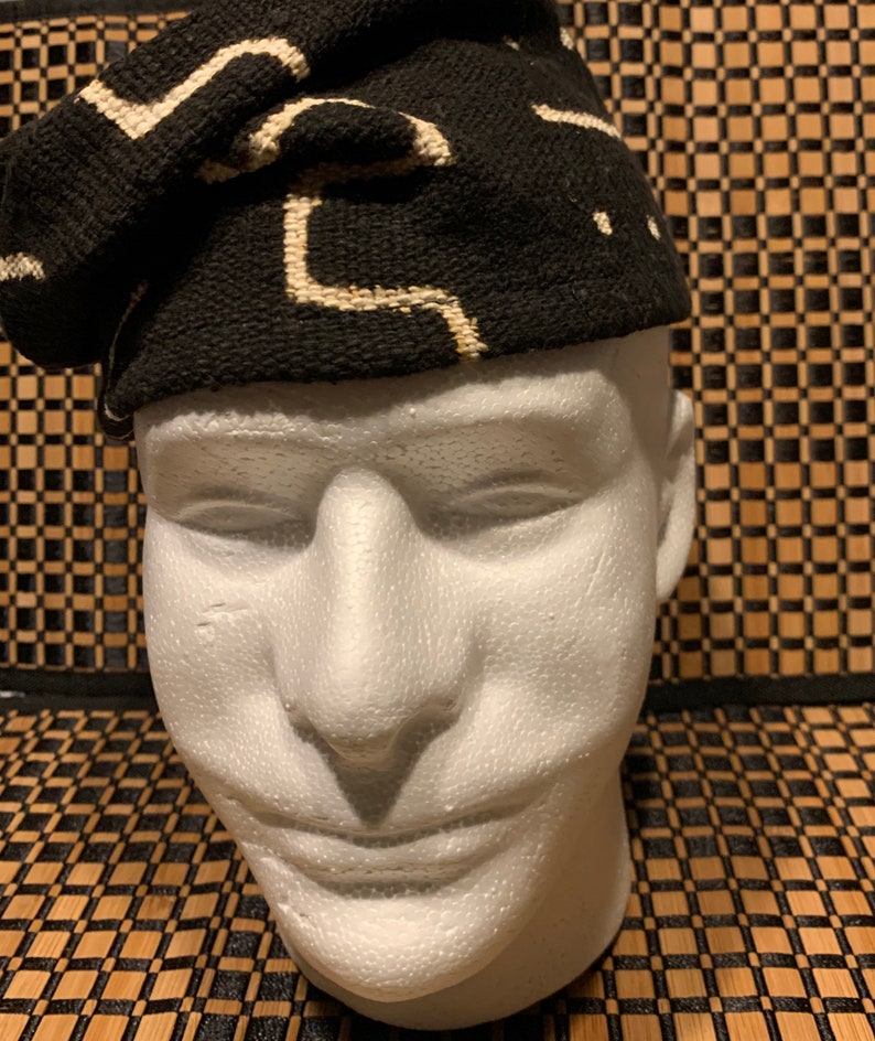 May include: A black and white patterned fabric hat. The hat is made of a woven fabric with a geometric design. The hat is folded and placed on a white mannequin head.