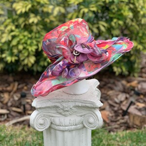 May include: A large, colorful hat with a pink, purple, and red floral design. The hat is made of a sheer fabric and has a wide brim. It is sitting on a white pedestal.