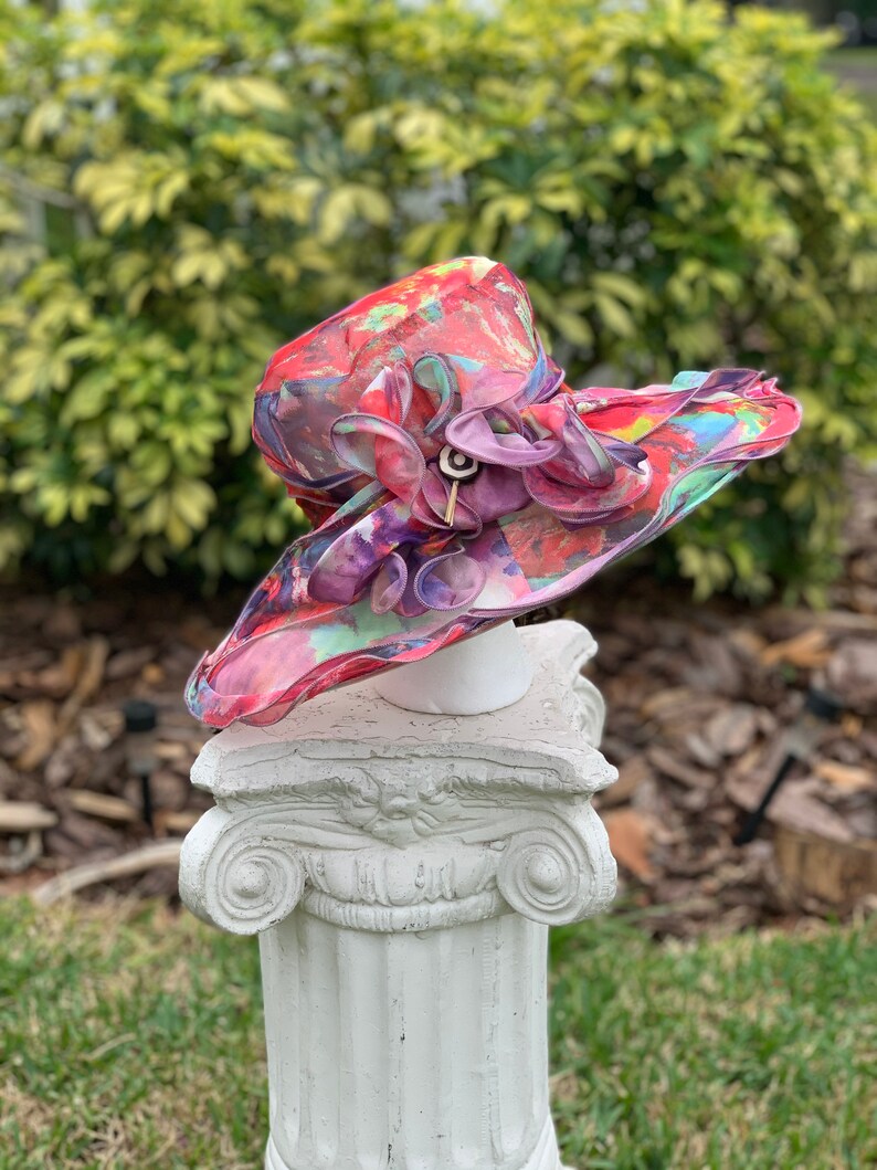 May include: A large, colorful hat with a pink, purple, and red floral design. The hat is made of a sheer fabric and has a wide brim. It is sitting on a white pedestal.