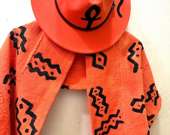 May include: An orange fedora hat with black decorative accents and an ankh symbol, paired with a matching orange scarf featuring a black geometric pattern. The hat and scarf are displayed against a white background.
