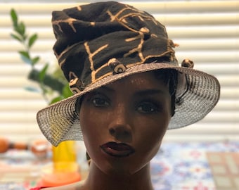 May include: A black and tan patterned hat with a wide brim. The hat has a decorative band and embellishments. The hat is displayed on a mannequin head, highlighting its design and style. The hat's brim is a silver colour.