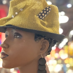 May include: A tan fedora-style hat with a black brim and a decorative polka-dot accent. The hat is adorned with three clear rhinestones. The mannequin is also wearing long, black, circular earrings.