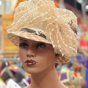 Op de afbeelding: Een beige strohoed met een grote witte bloem en een zwart lint. De hoed heeft een gaasontwerp en is versierd met kleine witte kralen.