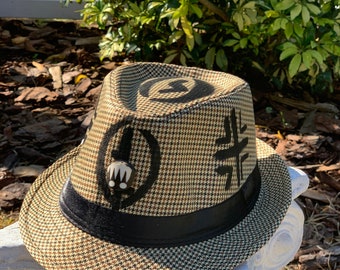 May include: A fedora hat with a houndstooth pattern in brown and tan, featuring black accents and decorative symbols. The hat has a black band and is set against a natural outdoor backdrop.