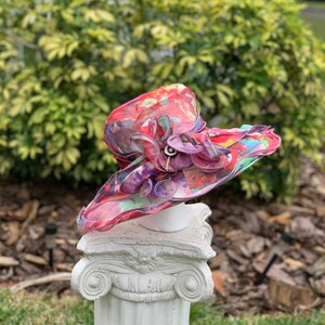 May include: A colorful floral hat with a large brim and a pink and purple flower accent. The hat is displayed on a white pedestal.