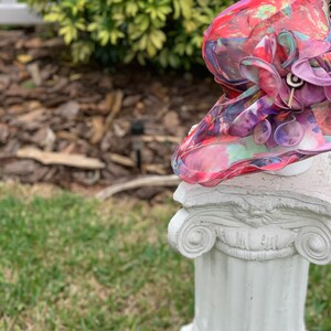 May include: A colorful floral hat with a large purple flower on top, sitting on a white pedestal.