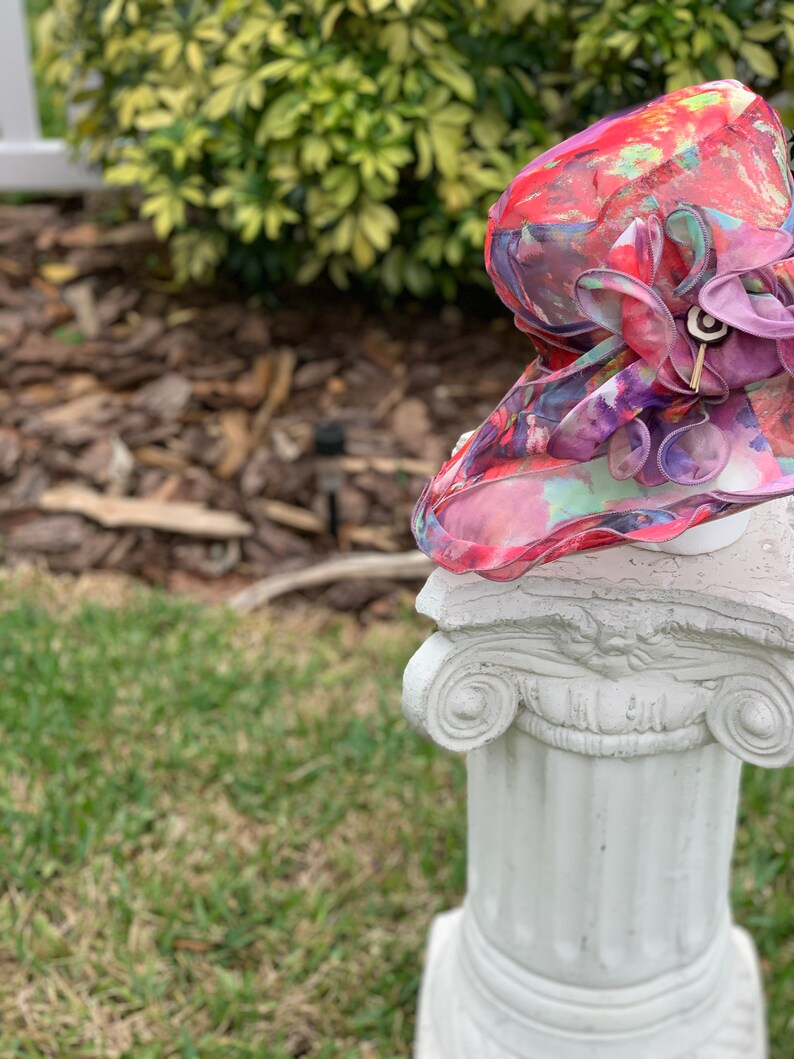 May include: A colorful floral hat with a large purple flower on top, sitting on a white pedestal.