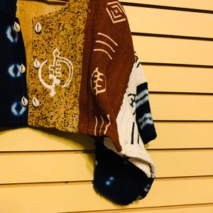 May include: A brown, white, and blue patterned cardigan with a button closure and a white design on the front. The cardigan is made of a woven fabric and has a relaxed fit.