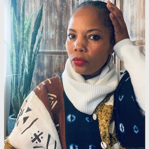 May include: A woman wearing a white turtleneck sweater and a multi-colored patterned cardigan with shell buttons. The cardigan has a brown, white, and blue pattern with geometric shapes and eye designs.