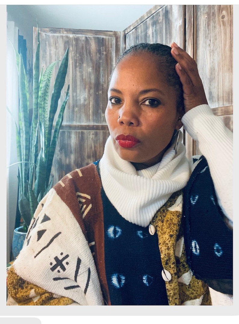 May include: A woman wearing a white turtleneck sweater and a multi-colored patterned cardigan with shell buttons. The cardigan has a brown, white, and blue pattern with geometric shapes and eye designs.