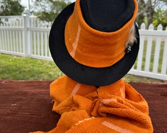 May include: A close-up of an orange and black fedora hat with a black band and a small feather accent. The hat is sitting on a brown surface.