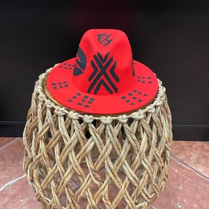 May include: A red fedora hat with black geometric designs. The hat is sitting on a woven wicker stool.