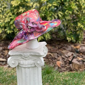 May include: A large, wide-brimmed hat with a colorful abstract print. The hat is made of a sheer fabric and has a large flower embellishment on the side. The hat is displayed on a white pedestal.