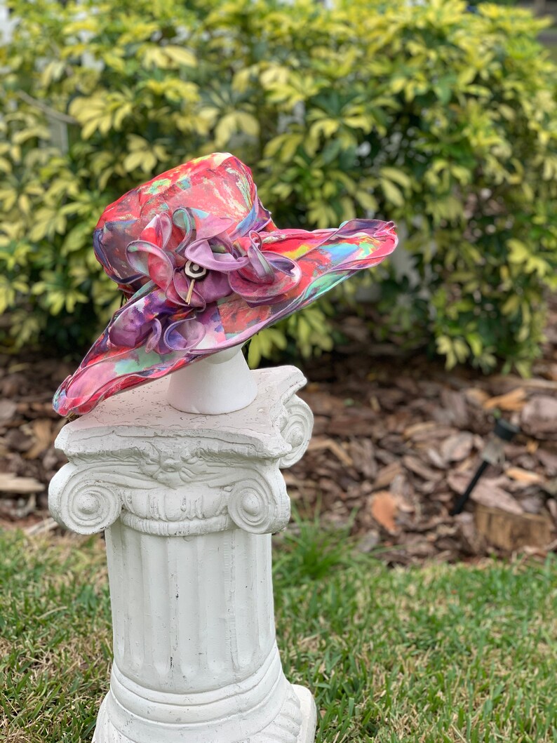 May include: A large, wide-brimmed hat with a colorful abstract print. The hat is made of a sheer fabric and has a large flower embellishment on the side. The hat is displayed on a white pedestal.