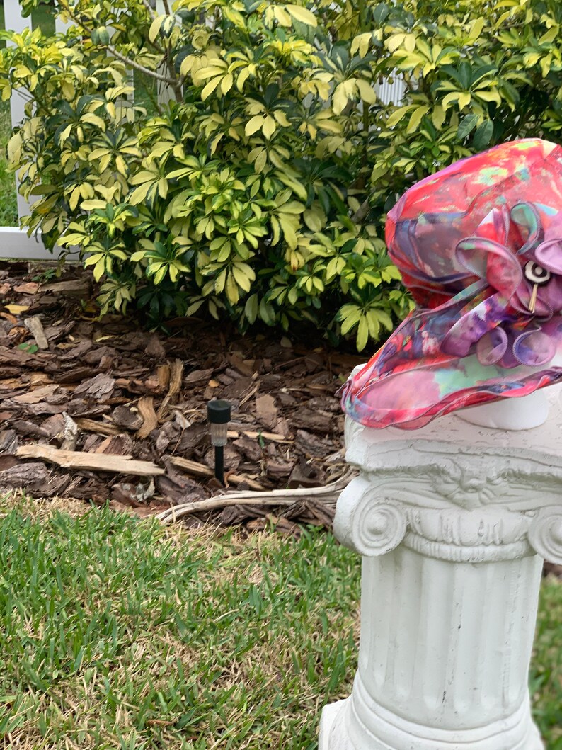 May include: A pink and red floral hat with a large flower accent sits on top of a white column.
