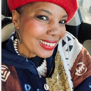 May include: A woman wearing a red beret, a blue and white patterned jacket, and a white beaded necklace. She is smiling and looking at the camera.