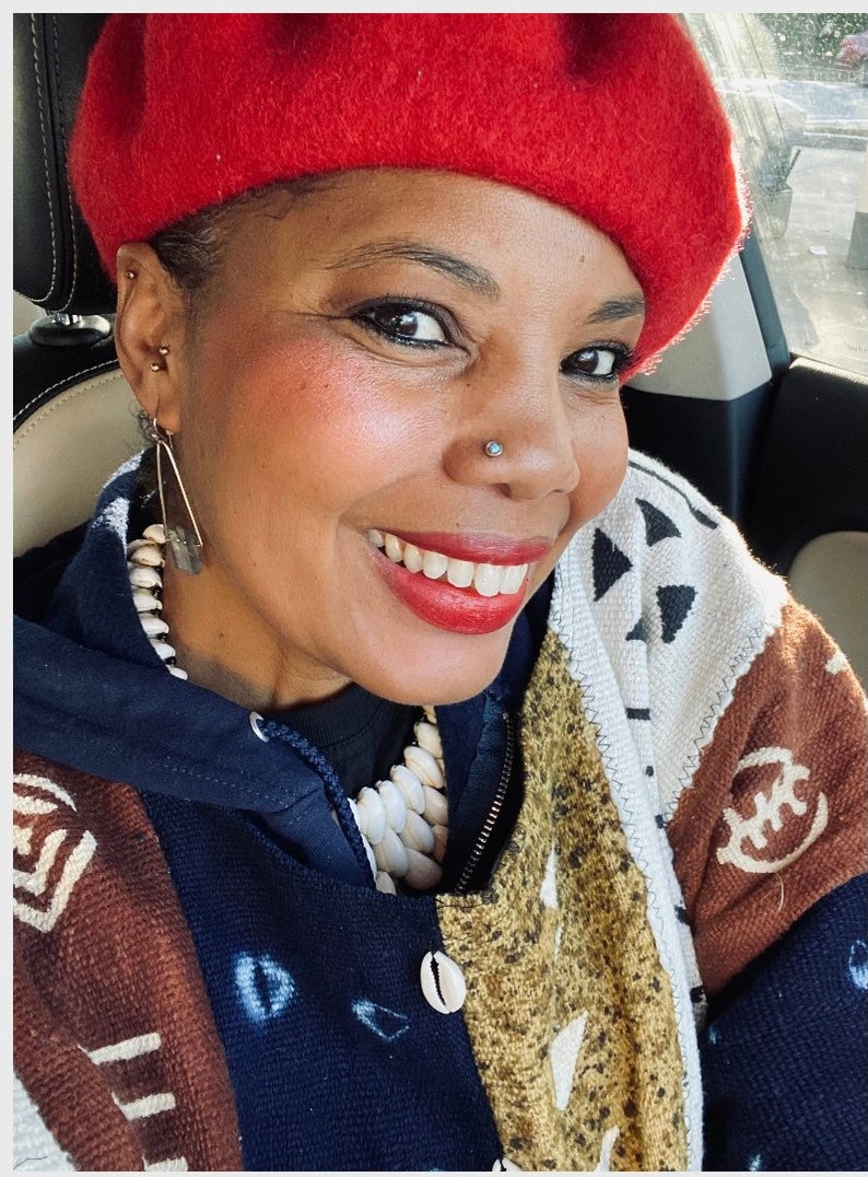May include: A woman wearing a red beret, a blue and white patterned jacket, and a white beaded necklace. She is smiling and looking at the camera.