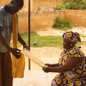 May include: A person in a blue shirt and brown pants is handing a white cloth to a person in a wheelchair. The person in the wheelchair is wearing a colorful dress and a headscarf. The wheelchair has a black seat and a black and silver frame.