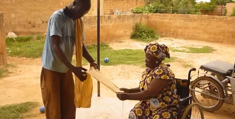 May include: A person in a blue shirt and brown pants is handing a white cloth to a person in a wheelchair. The person in the wheelchair is wearing a colorful dress and a headscarf. The wheelchair has a black seat and a black and silver frame.