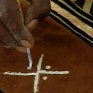May include: A person's hand using a tool to draw a white cross on a brown surface with black and white stripes.