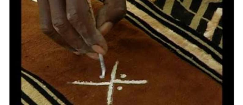 May include: A person's hand using a tool to draw a white cross on a brown surface with black and white stripes.
