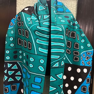 Peut inclure: Écharpe turquoise et bleu sarcelle avec un motif géométrique. L'écharpe présente un motif répétitif de carrés, de cercles et de triangles en noir, blanc et bleu. L'écharpe est drapée sur une surface noire.