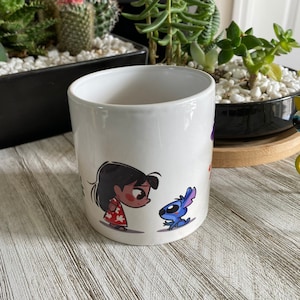 Puede incluir: Taza de cerámica blanca con una imagen de dibujos animados de una niña con cabello oscuro que lleva un vestido rojo y blanco y un personaje alienígena azul con orejas grandes. La niña está mirando al personaje alienígena.