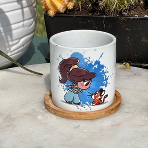 Peut inclure: Un pot en céramique blanche avec un motif aquarelle bleu et blanc représentant une princesse de dessin animé et un tigre. Le pot est posé sur un dessous-de-plat en bois.