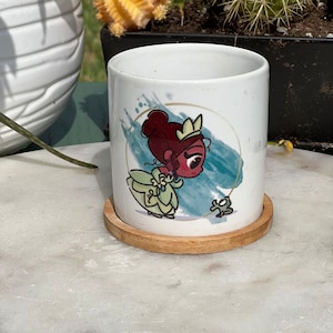 May include: A white ceramic mug with a cartoon illustration of a princess with a crown and a frog. The mug is sitting on a wooden coaster.