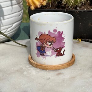May include: White ceramic mug with a cartoon illustration of a girl with brown hair holding a small stuffed animal and a red cat looking at her. The mug is on a wooden coaster.