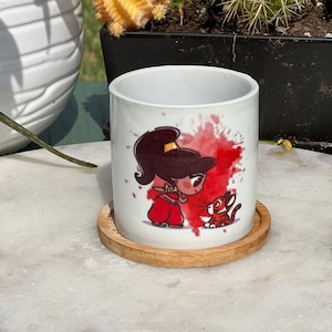 Peut inclure: Une tasse en céramique blanche avec une image de dessin animé d'une femme en robe rouge et d'un tigre. La tasse est posée sur un dessous de verre en bois.