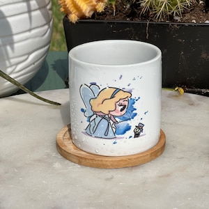 Könnte beinhalten: Eine weiße Keramiktasse mit einem blauen und violetten Aquarelldesign, das eine Comic-Fee mit blonden Haaren und einem blauen Kleid zeigt. Die Tasse steht auf einem Holz-Untersetzer.