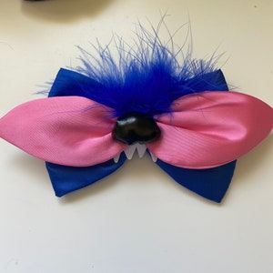 May include: A pink and blue bow with a black nose and white teeth, topped with blue feathers.