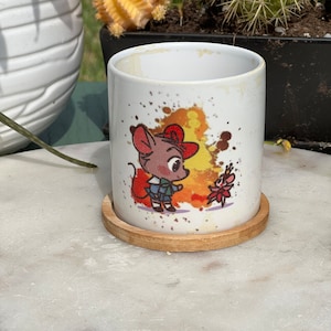 May include: White ceramic mug with a cartoon mouse wearing a red hat and a blue shirt. The mug has a watercolor-style background with orange, yellow, and pink colors. The mug is sitting on a wooden coaster.