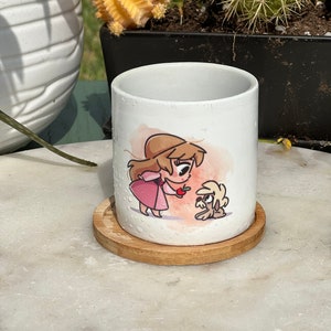 May include: White ceramic mug with a cartoon illustration of a girl in a pink dress giving an apple to a small dog. The mug is on a wooden coaster.
