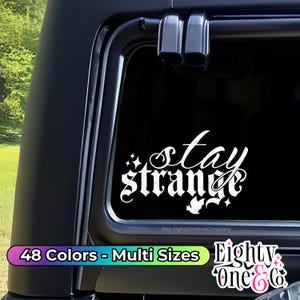 May include: A white vinyl decal with the text "Stay Strange" in a gothic font. The decal is on a black car window. The text is surrounded by small stars and a bird.