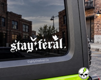 May include: A white vinyl decal applied to a vehicle window featuring the text stay feral in a gothic font, accented with small star shapes and two bat silhouettes. The text EightyOneAnd.Co is printed below the main decal design.