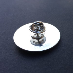 May include: The back of a silver pin with a circular backing and a safety pin closure.