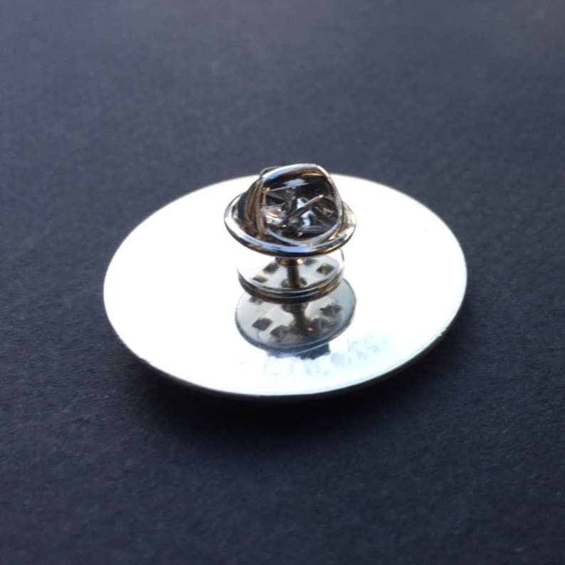 May include: The back of a silver pin with a circular backing and a safety pin closure.