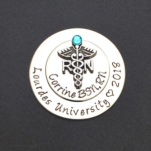 May include: Silver round charm with a blue gem, a caduceus, and the letters "RN" engraved. The text "Corrine BSN, RN" and "Our Lady of Lourdes University" is engraved around the edge. The date "2018" is engraved on the bottom.