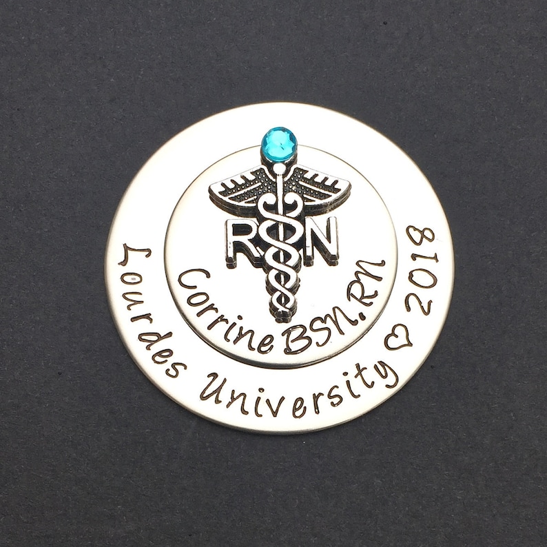 May include: Silver round charm with a blue gem, a caduceus, and the letters "RN" engraved. The text "Corrine BSN, RN" and "Our Lady of Lourdes University" is engraved around the edge. The date "2018" is engraved on the bottom.